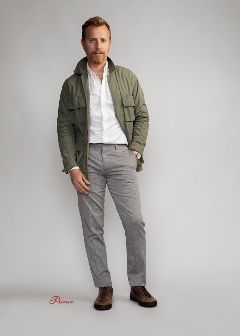 a man wearing a smart casual green field jacket with white button down shirt, grey linen pants, and brown sneakers