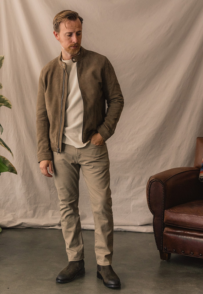 A man with short hair and a short beard is standing in front of a light-colored draped fabric backdrop. He is wearing a tan suede jacket, a white long-sleeve shirt, beige pants, and brown leather boots. He has one hand in his pocket and the other resting by his side. 