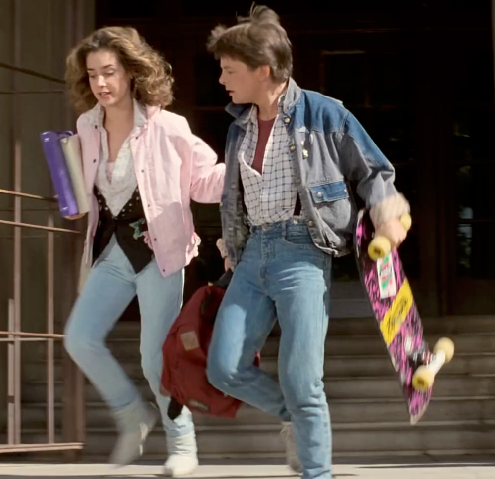 Screenshot from Back to the Future featuring Jenifer and Marty in light wash jeans. The woman has curly hair and wears a light pink jacket over a white shirt with light blue jeans, holding books in her arm. The man has short hair and wears a denim jacket over a checkered shirt with blue jeans, carrying a red backpack and a skateboard with a colorful design.