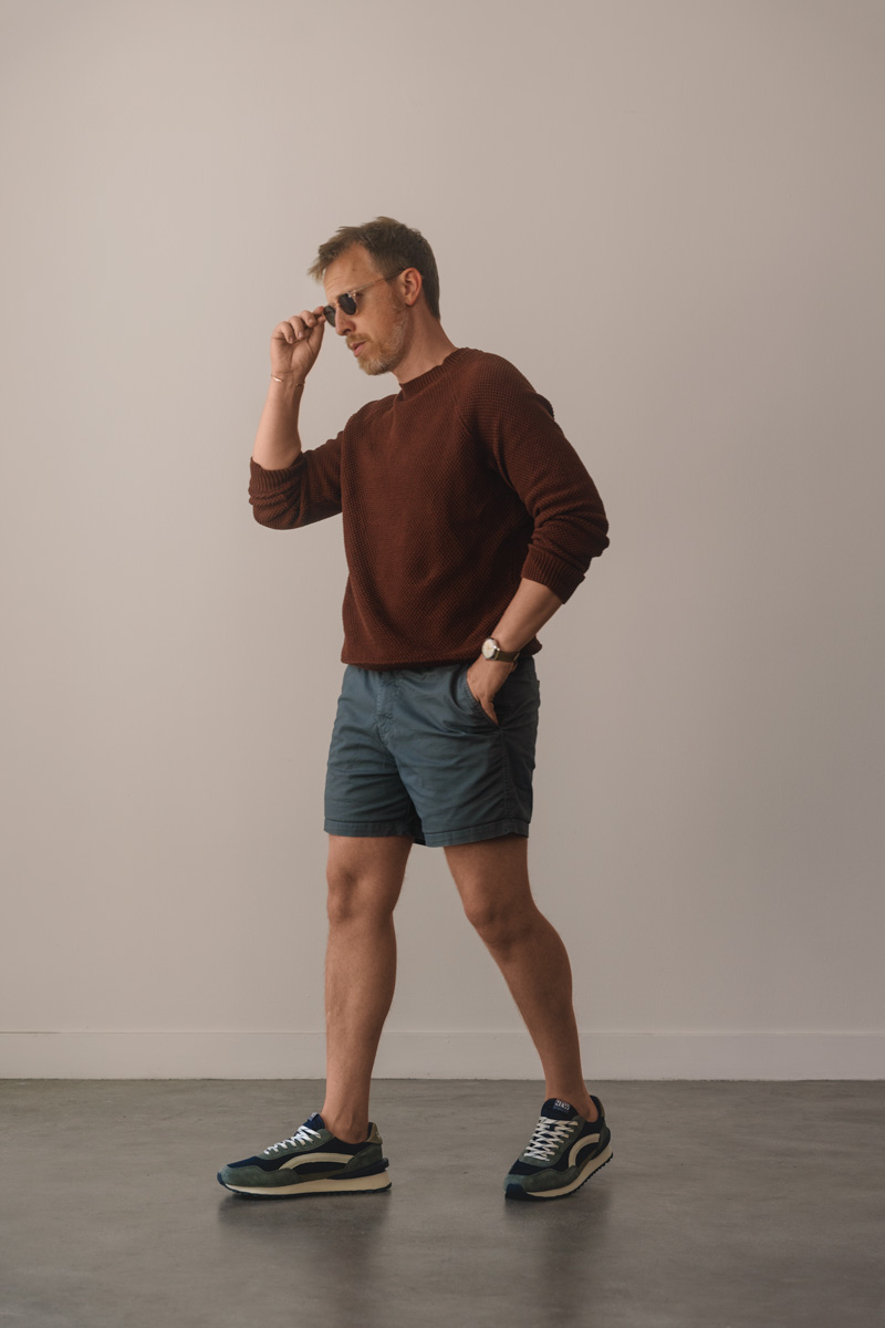men's summer outfit idea with a red sweater, light blue shorts, and green and blue running sneakers