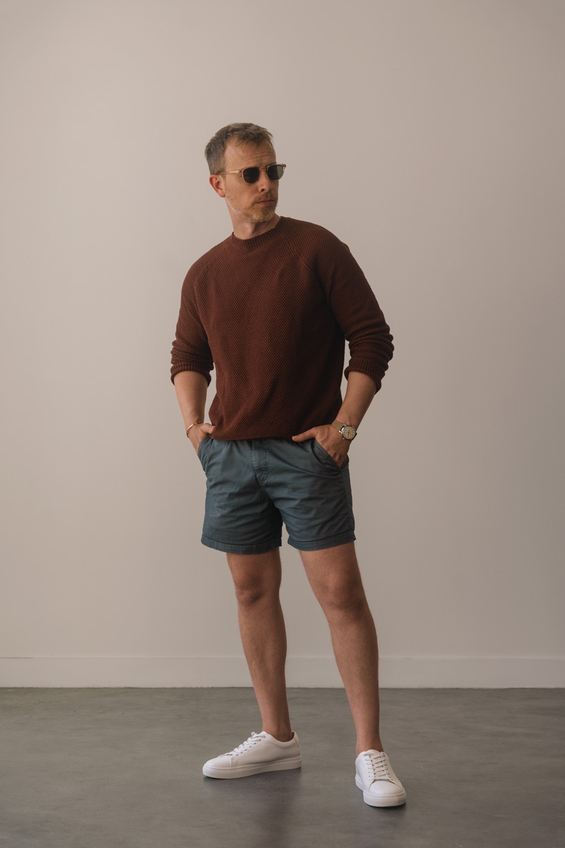 men's summer outfit idea with a red sweater, light blue shorts, and white minimalist sneakers
